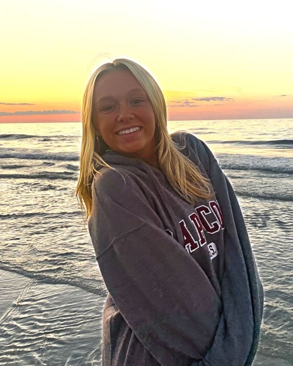 Ruby wearing a gray sweatshirt in front of sunset with the beach in the background.