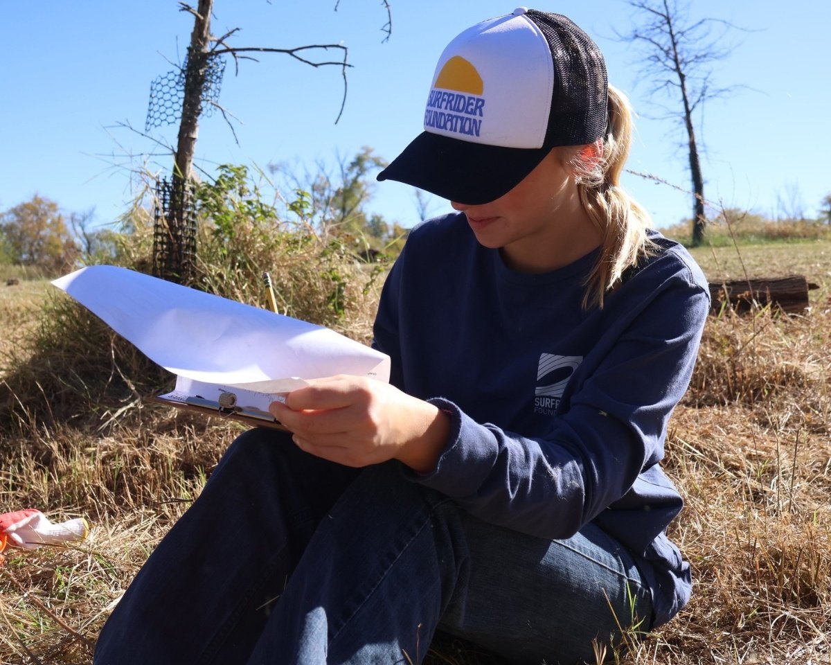 Ruby in the field testing water quality, wearing a hat and holding a notepad.