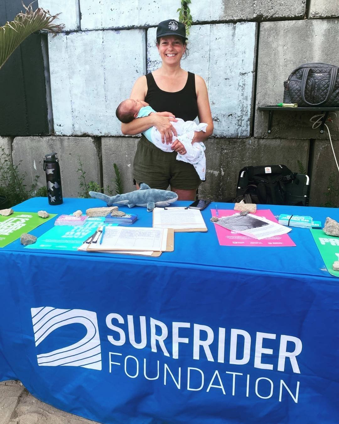 Corinne holds her baby while standing at a table with a blue Surfrider tablecloth.